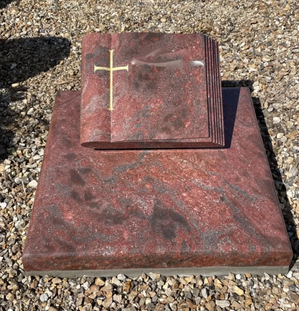 Plaque funéraire en granit rouge en forme de livre ouvert avec croix dorée.